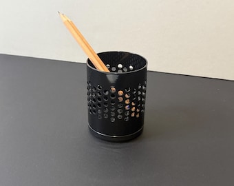 May include: A black cylindrical pencil holder with a perforated design. A wooden pencil is placed inside the holder, angled upwards. The pencil holder sits on a dark surface, with a light-colored wall in the background.