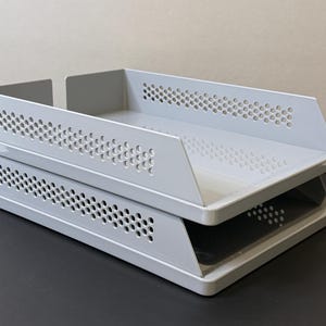 Op de afbeelding: Twee niveaus grijze metalen bureau-organizer. Elke lade heeft een geperforeerd ontwerp langs de zijkanten. De laden zijn op elkaar gestapeld. De organizer is ontworpen om papieren en documenten in op te bergen.