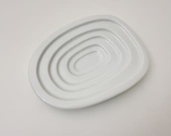 May include: A white, ceramic soap dish with a unique, layered design. The dish features a series of concentric, oval-shaped ridges, creating a textured surface. The overall shape is an elongated oval, perfect for holding a bar of soap.