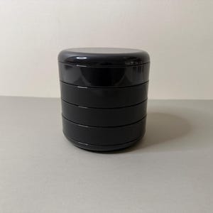 May include: A black, stackable container with a rounded top and four visible sections. The container is made of a glossy material and sits on a light grey surface. The background is a plain, off-white wall.