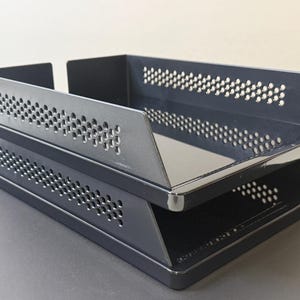 May include: A two-tiered, dark blue metal desk organiser. The trays have a perforated design along the sides and angled edges. The organiser is designed to hold papers and documents.