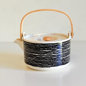 Puede incluir: Una tetera de cerámica blanca con un patrón de puntos negros y un asa de madera. La tapa tiene un pomo de madera. El pico es blanco. La tetera está diseñada para preparar té.