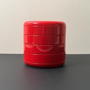 Może przedstawiać: Stapialny, czerwony pojemnik z błyszczącym wykończeniem. Pojemnik składa się z czterech oddzielnych, okrągłych sekcji, które pasują do siebie. Produkt stoi na ciemnej powierzchni z jasną ścianą w tle.