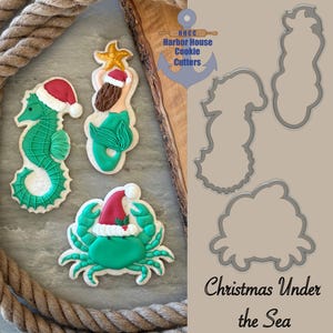 May include: Christmas-themed cookies shaped like a seahorse, mermaid, and crab, all wearing Santa hats. The cookies are decorated with green, red, and white icing. Cookie cutters in the shape of the cookies are also shown. The text "Christmas Under the Sea" is visible.