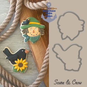 May include: Scarecrow and crow-themed cookies with a sunflower, displayed on a wooden surface with rope. The cookies are decorated with green, yellow, black, and brown icing. Two metal cookie cutters are also visible. The text "Scare & Crow" is at the bottom.