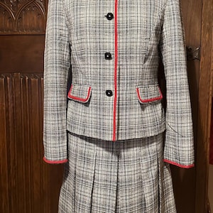 May include: A two-piece plaid suit featuring a jacket and a pleated skirt. The jacket has a round neckline, red trim, and black buttons. The skirt is knee-length. The suit is displayed on a mannequin.