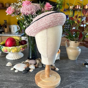 Op de afbeelding: Een lichtroze gehaakte baret met een zwarte rand, tentoongesteld op een mannequinhoofd. De hoed is gemaakt van getextureerd garen en heeft een klassieke baretvorm. De achtergrond omvat bloemen en een decoratieve fruitschaal.