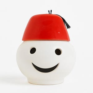 Può includere: Un vaso di ceramica bianco con un design a forma di viso sorridente, con fori neri per gli occhi e una bocca curva. È sormontato da un coperchio rosso brillante in stile fez con un nappa nera. Il vaso ha un aspetto stravagante e decorativo.