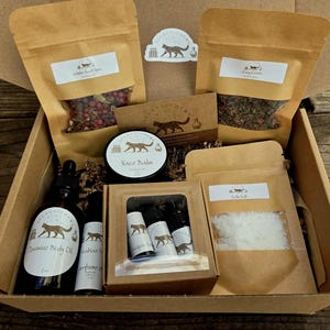 Puede incluir: Una caja de regalo llena de varios productos de belleza y bienestar. La caja contiene varias bolsas de papel marrón con etiquetas, botellas pequeñas, un tarro de bálsamo facial y aceite corporal de jazmín. Los productos están dispuestos sobre una superficie de madera.