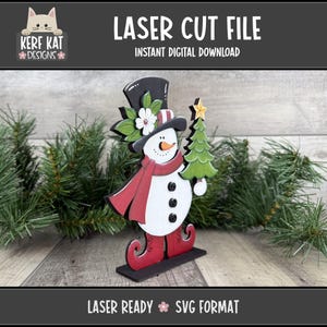 May include: A laser-cut snowman decoration with a black top hat, red scarf, and holding a green Christmas tree with a yellow star. The snowman has a carrot nose and black button eyes. The text "LASER CUT FILE" and "INSTANT DIGITAL DOWNLOAD" are at the top.