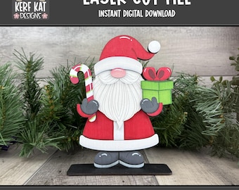 May include: A laser-cut Santa Claus figure, holding a candy cane and a green gift box with a red bow. The figure is red, white, and gray, with the text "LASER CUT FILE" and "SVG FORMAT".