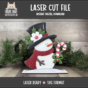 May include: A laser cut file of a snowman with a black top hat, red scarf, and candy cane. The snowman has a carrot nose and a flower on its hat. The text "LASER CUT FILE" and "INSTANT DIGITAL DOWNLOAD" are displayed.
