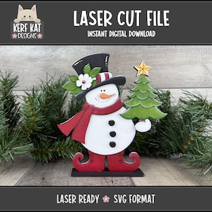 May include: A laser-cut snowman decoration with a black top hat, red scarf, and holding a green Christmas tree. The snowman has a carrot nose, black button eyes, and a smiling mouth. The text "LASER CUT FILE" and "INSTANT DIGITAL DOWNLOAD" are displayed.
