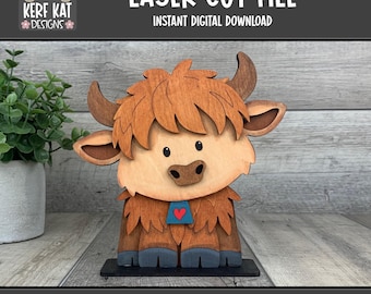 May include: A wooden laser cut file of a Highland cow. The cow has light brown fur, dark brown horns, and a blue heart-shaped pendant. The text "LASER CUT FILE" and "INSTANT DIGITAL DOWNLOAD" are at the top. The bottom text reads "LASER READY SVG FORMAT".