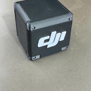 Könnte beinhalten: Ein schwarzer, würfelförmiger Koffer mit abgerundeten Ecken und einem weißen Logo auf der Vorderseite. Das Logo ist eine stilisierte Darstellung der Buchstaben "DJI". Der Koffer hat kleine, silberfarbene Befestigungselemente an jeder Ecke.