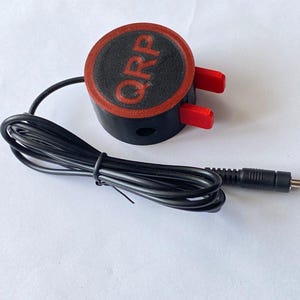 Könnte beinhalten: Ein schwarzer und roter Audioadapter mit einem schwarzen Kabel und einem 3,5-mm-Stecker. Der Adapter trägt die Aufschrift "QRP".