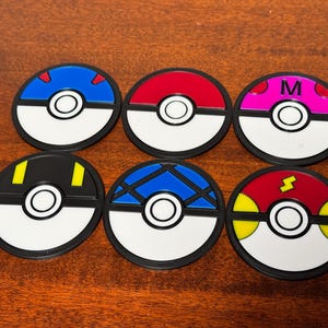 Puede incluir: Seis posavasos redondos, negros y blancos de estilo Poké Ball con diferentes acentos de color. Los posavasos presentan un acento rojo, azul, rosa, amarillo, negro y azul.