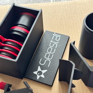 May include: A selection of black and red astronomy equipment components. The image shows a rectangular box with red and black circular pieces, a cylindrical tube, and a plate with the word "Seestar" in white.