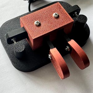 Pode incluir: Um dispositivo mecânico com uma base preta e uma seção superior texturizada, marrom-avermelhada, fixada com parafusos prateados. Duas alavancas marrom-avermelhadas se estendem do dispositivo, sugerindo um pedal ou mecanismo de controle. O design é industrial e funcional.