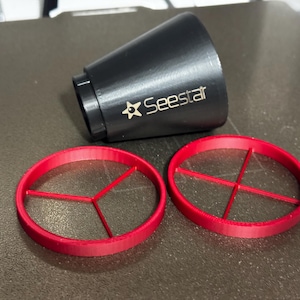 Może przedstawiać: Czarne, stożkowe urządzenie z napisem "Seestar" i złotym logo gwiazdy. Obecne są również dwa czerwone, okrągłe filtry z krzyżową strukturą nośną. Przedmioty te są prawdopodobnie związane z astronomią lub optyką.