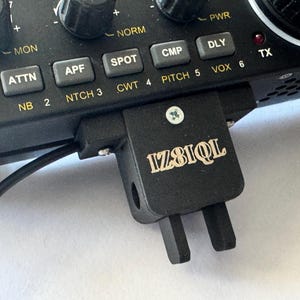 Peut inclure: Gros plan sur un émetteur-récepteur radio noir avec un câble de microphone noir attaché. La radio possède une variété de boutons et de molettes, notamment un bouton de volume, un bouton d'alimentation et une prise microphone. La radio possède également un petit appareil rectangulaire noir qui lui est attaché avec le texte "IZ8IQL" imprimé dessus.