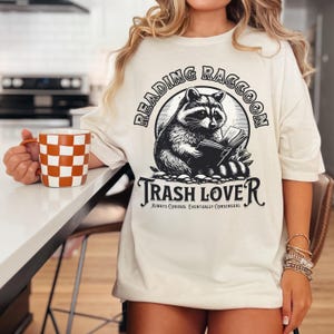 Puede incluir: Camiseta color crema con una ilustraci&oacute;n en blanco y negro de un mapache leyendo un libro. El texto "READING RACCOON" est&aacute; arqueado sobre el mapache, y "TRASH LOVER" est&aacute; debajo. Tambi&eacute;n se ve una taza con un patr&oacute;n de cuadros naranja y blanco.