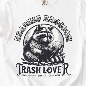 Puede incluir: Camiseta blanca con una ilustraci&oacute;n en blanco y negro de un mapache leyendo un libro. El texto "READING RACCOON" est&aacute; arqueado sobre el mapache, y "TRASH LOVER" est&aacute; debajo. Tambi&eacute;n se incluye la frase "ALWAYS CURIOUS. EVENTUALLY CONSENSUAL".