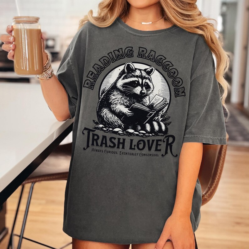 Puede incluir: Camiseta gris oscuro con una ilustraci&oacute;n en blanco y negro de un mapache leyendo un libro. El texto en la camiseta dice "Reading Raccoon" encima del mapache y "Trash Lover" debajo, con el texto adicional "Always Curious, Eventually Consensual".