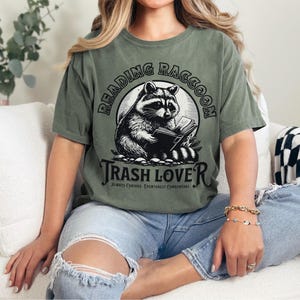 Puede incluir: Camiseta verde oliva con una ilustraci&oacute;n en blanco y negro de un mapache leyendo un libro. El texto "READING RACCOON" est&aacute; arqueado sobre el mapache, con "TRASH LOVER" debajo. La frase "ALWAYS CURIOUS. EVENTUALLY CONSENSUAL" est&aacute; impresa debajo.