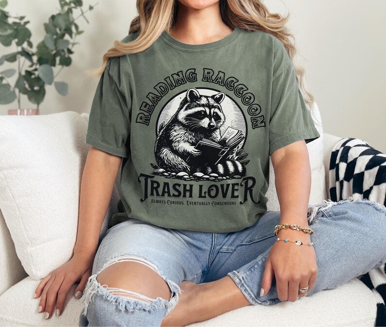 Puede incluir: Camiseta verde oliva con una ilustraci&oacute;n en blanco y negro de un mapache leyendo un libro. El texto "READING RACCOON" est&aacute; arqueado sobre el mapache, con "TRASH LOVER" debajo. La frase "ALWAYS CURIOUS. EVENTUALLY CONSENSUAL" est&aacute; impresa debajo.