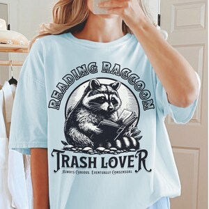 Puede incluir: Camiseta azul claro con una ilustraci&oacute;n en blanco y negro de un mapache leyendo un libro. El texto "READING RACCOON" est&aacute; encima del mapache, y "TRASH LOVE" est&aacute; debajo. La camiseta tambi&eacute;n incluye el texto "ALWAYS CURIOUS. EVENTUALLY CONSENSUAL."