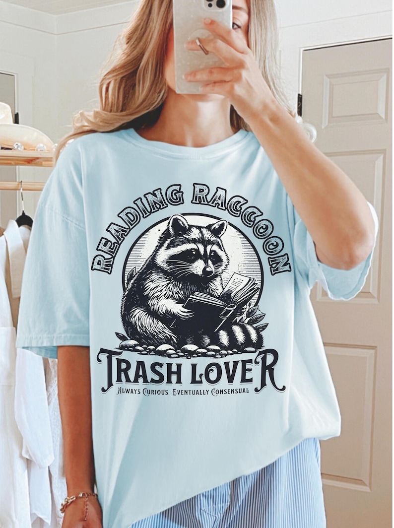 Puede incluir: Camiseta azul claro con una ilustraci&oacute;n en blanco y negro de un mapache leyendo un libro. El texto "READING RACCOON" est&aacute; encima del mapache, y "TRASH LOVE" est&aacute; debajo. La camiseta tambi&eacute;n incluye el texto "ALWAYS CURIOUS. EVENTUALLY CONSENSUAL."