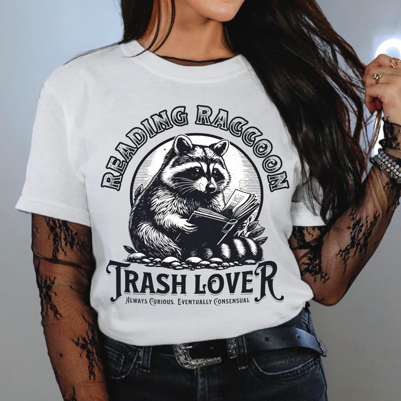 Puede incluir: Camiseta blanca con una ilustraci&oacute;n en blanco y negro de un mapache leyendo un libro. El texto "Reading Raccoon" est&aacute; encima del mapache, y "Trash Lover" est&aacute; debajo. Tambi&eacute;n se incluye la frase "Always Curious. Eventually Consensual".