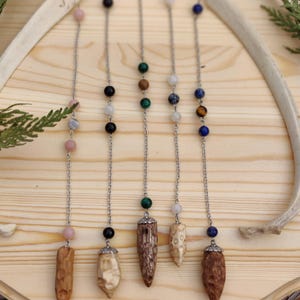 May include: Five pendulums with silver chains and various gemstone beads. Each pendulum has a different wooden or stone point. The gemstones include pink, black, green, white, blue, and tiger's eye. The pendulums are displayed on a wooden surface.