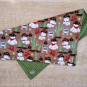 May include: A brown and green pet bandana with a cowboy ghost pattern. The bandana features a repeating design of ghosts wearing cowboy hats, cacti, and wanted posters. The reverse side is green with white polka dots. Grommets are visible.