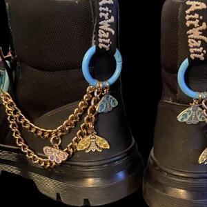 Puede incluir: Botas negras con cadenas doradas y anillos azules unidos a los cordones. Las cadenas tienen pequeños colgantes de mariposa.