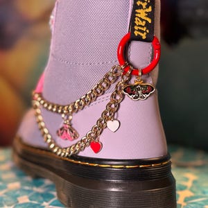 May include: A lavender boot with a black sole and gold trim, adorned with a gold chain and charms. Charms include a moth, a dress, and heart shapes. The boot features a red ring and the text "AirWair" in yellow.