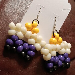 May include: A pair of beaded earrings with a yellow, white, and purple color scheme. The earrings are made of small, round beads and are attached to silver hooks.
