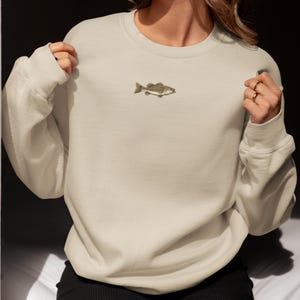 May include: A cream-colored sweatshirt featuring a small embroidered fish design on the chest. The fish is detailed with shades of brown and tan. The sweatshirt has long sleeves and a crew neck.