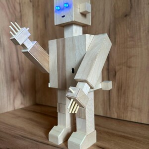 Può includere: Scultura di robot in legno con finitura in legno chiaro. Il robot ha un design a blocchi con occhi a LED blu e braccia articolate. Le mani del robot hanno estensioni di legno a forma di dita. Il robot è su una superficie di legno.