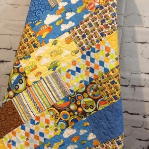 May include: A patchwork quilt with a diagonal pattern. The quilt is made of various fabrics with different designs, including airplanes, trucks, and circles. The quilt is in shades of blue, yellow, orange, green, and brown.