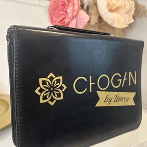 Könnte beinhalten: Schwarze Ledertasche mit goldenem Blumendesign und dem Text "CHOGAN by Ümra". Die Tasche hat einen Reißverschluss und einen Griff. Der Hintergrund umfasst Blumen.