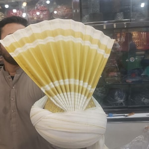Peut inclure: Un turban blanc et doré avec un motif en forme d'éventail. Le turban a une base arrondie et un dessus plissé en forme d'éventail avec des rayures blanches et dorées alternées. La base a des broderies dorées.