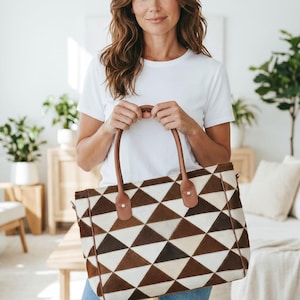 May include: A rectangular tote bag featuring a geometric pattern of alternating brown and cream-colored triangular patches. The bag has two brown leather handles attached with silver-toned metal rivets at the top edge of the bag.