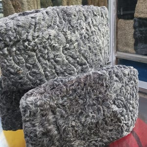 May include: Two grey fur hats with a textured, curly pattern. The hats are stacked, showing the detail of the fur. The hats appear to be made of a soft, warm material, ideal for cold weather. The hats are likely to be worn in winter.