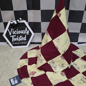 May include: A burgundy and cream-coloured wizard hat with a wide brim. The hat features a patchwork design with burgundy squares and cream-coloured squares printed with images of letters, books, and the number 9 3/4. The hat is next to a sign that says "Viciously Twisted Creations."