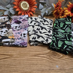 May include: Set of eight Halloween-themed fabric napkins featuring various spooky designs, including skulls, bats, bones, and flowers. The napkins are black, green, white, and pink.