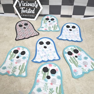 May include: A collection of handmade ghost-shaped fabric coasters. Some feature a cactus and floral print, others have a patterned design, and some have a starfish pattern. Each coaster has a black face and a contrasting border. The sign in the background reads "Viciously Twisted Creations."