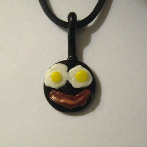 May include: A black glass frying pan pendant necklace with two white eggs and a brown bacon strip. The necklace is on a black cord.