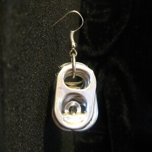 May include: A single silver-colored earring made from a recycled soda can tab. The earring features a hook and a circular ring connecting to the tab. The tab has a rounded rectangular shape with a raised center. The background is a dark, textured fabric.
